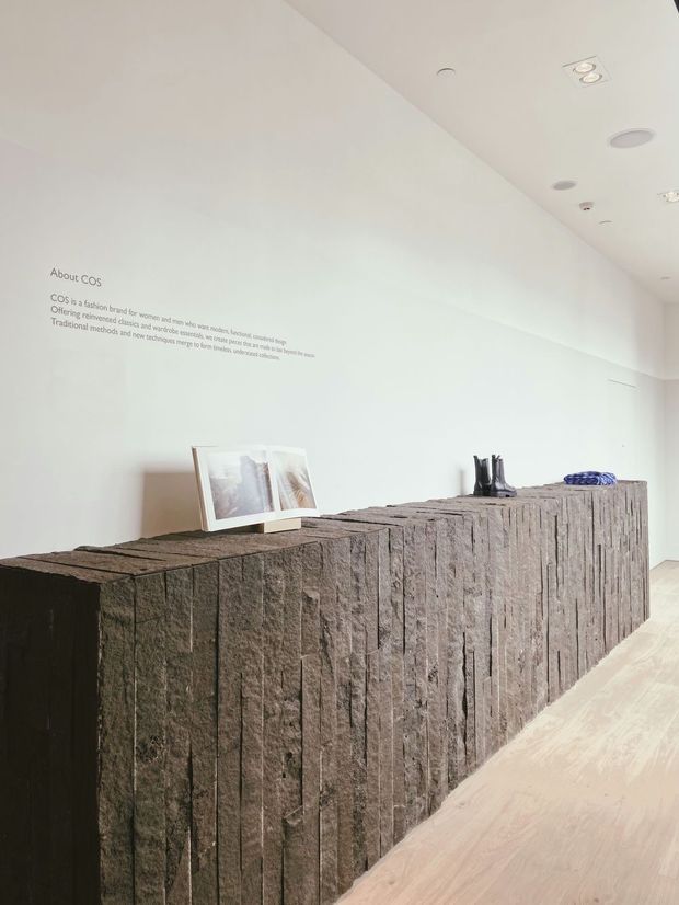 Minimalist retail or gallery-like interior with a long, sculptural dark stone reception or display counter featuring deeply textured vertical faces. The space has white walls, pale timber flooring, recessed ceiling lighting, sparse product displays, and understated wall text introducing COS.