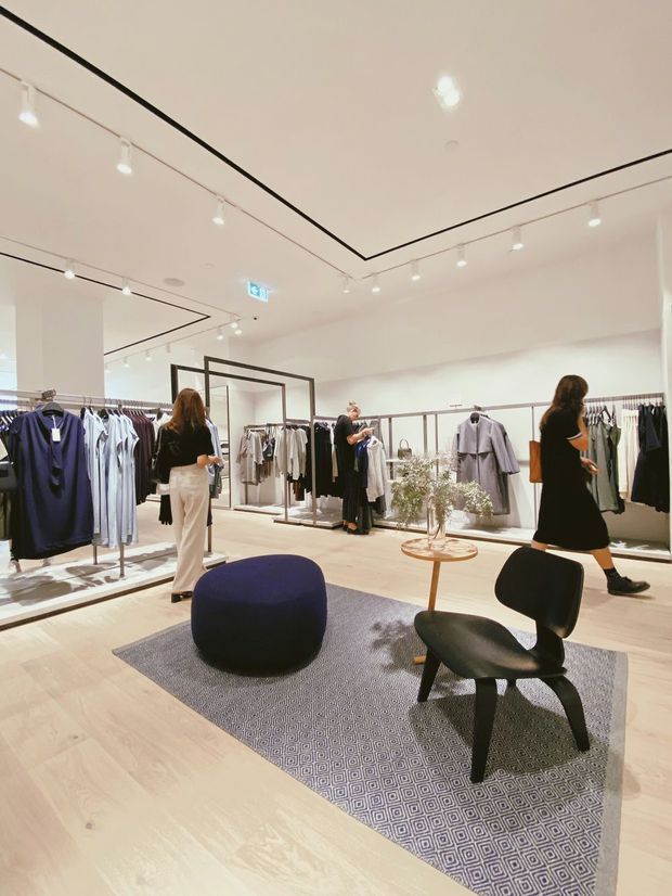 Bright contemporary clothing retail interior with pale timber flooring, white walls and ceiling, black linear ceiling detailing, track lighting, perimeter clothing racks, central display fixtures, a patterned area rug, and sculptural lounge seating. Shoppers and staff are present within the open, minimalist showroom.