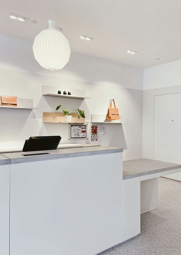 Bright minimalist reception or service counter area with white built-in cabinetry and open shelving, a pale grey stone-like countertop, integrated work surface, laptop, small decor, and a large sculptural white pendant light. The space has recessed ceiling lights, a closed white door, and a light grey carpeted floor.