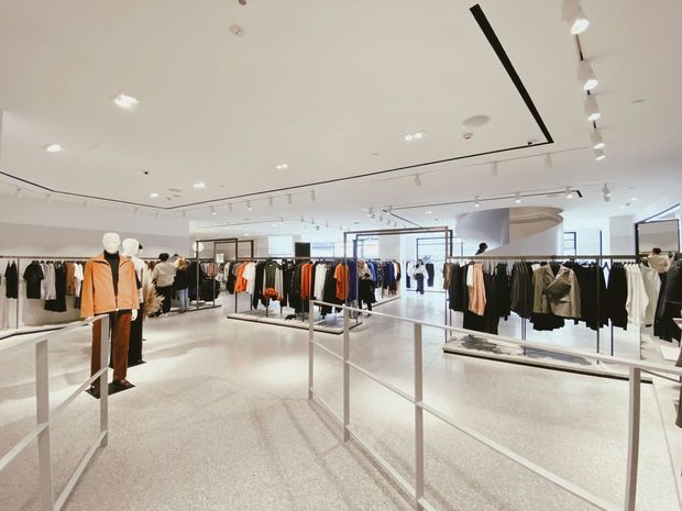 Bright contemporary clothing retail interior with extensive freestanding garment racks, mannequins, display tables, and track lighting. The open-plan store has a white ceiling, polished light terrazzo-like flooring, and a restrained white and black fit-out with clothing merchandise throughout.