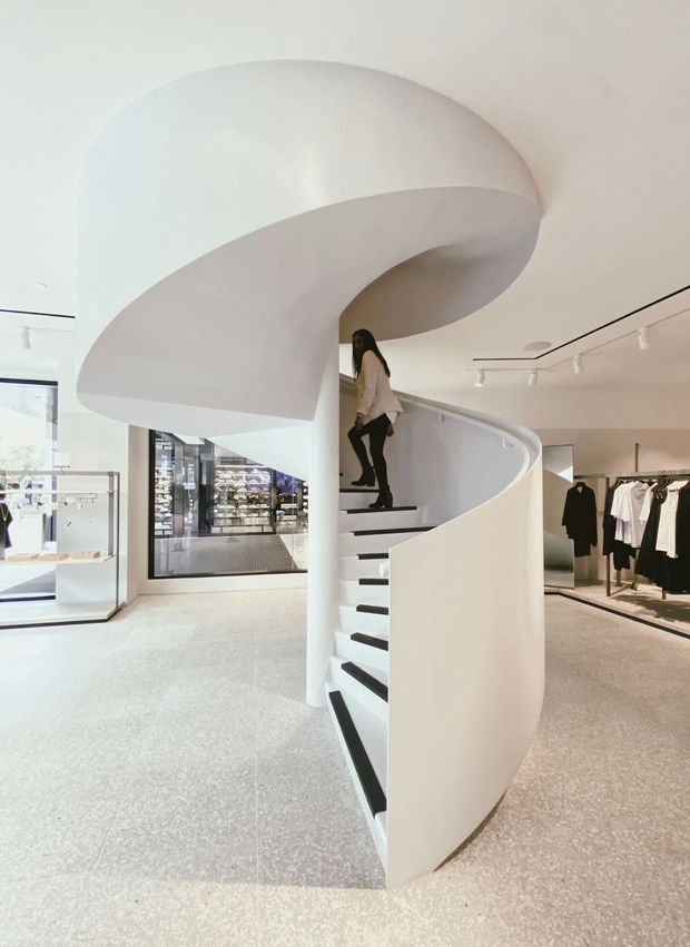 Bright minimalist retail interior featuring a dramatic sculptural white spiral staircase with black treads, curved balustrade, and a sweeping sculpted ceiling canopy. Clothing racks and glass display cases surround the staircase, with track lighting and polished light flooring creating an airy contemporary fashion-store setting.