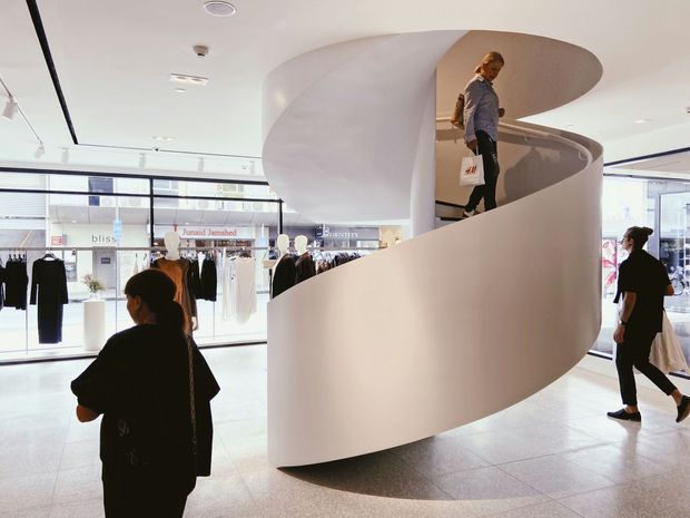 Bright contemporary commercial retail interior in a shopping centre, dominated by a sculptural white curved ribbon staircase with an open central void. Full-height glazing, track lighting, pale tiled flooring, clothing displays, and shoppers surround the staircase.