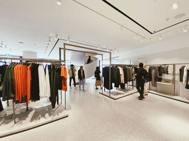Bright, spacious contemporary clothing retail store with minimalist white interior finishes, polished light flooring, ceiling track lighting, and multiple freestanding metal garment racks displaying apparel. A framed central display structure and built-in shelving define the open commercial layout.