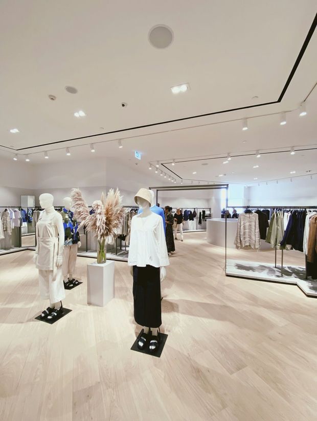 Bright contemporary fashion retail store interior with pale timber flooring, white walls and ceiling, extensive track lighting, clothing racks, display plinths, mannequins, and large mirrors. The open plan layout has a clean, minimalist presentation with neutral clothing displays and a central floral arrangement.