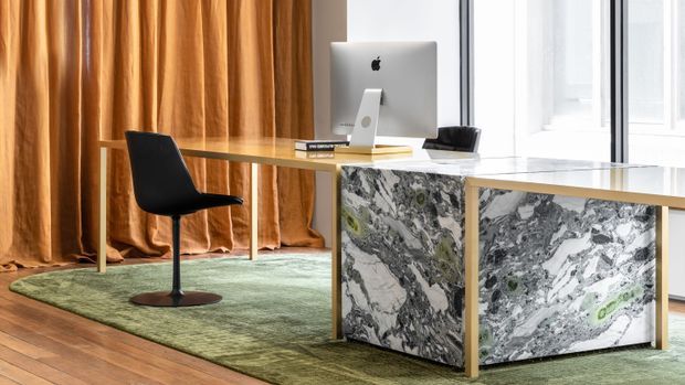 A refined home office with a slim glass-top desk and sculptural marble-clad drawer pedestal in white, grey, and green veining. A black modern desk chair, desktop computer, books, green area rug, warm timber flooring, full-height ochre curtains, and bright glazing complete the space.