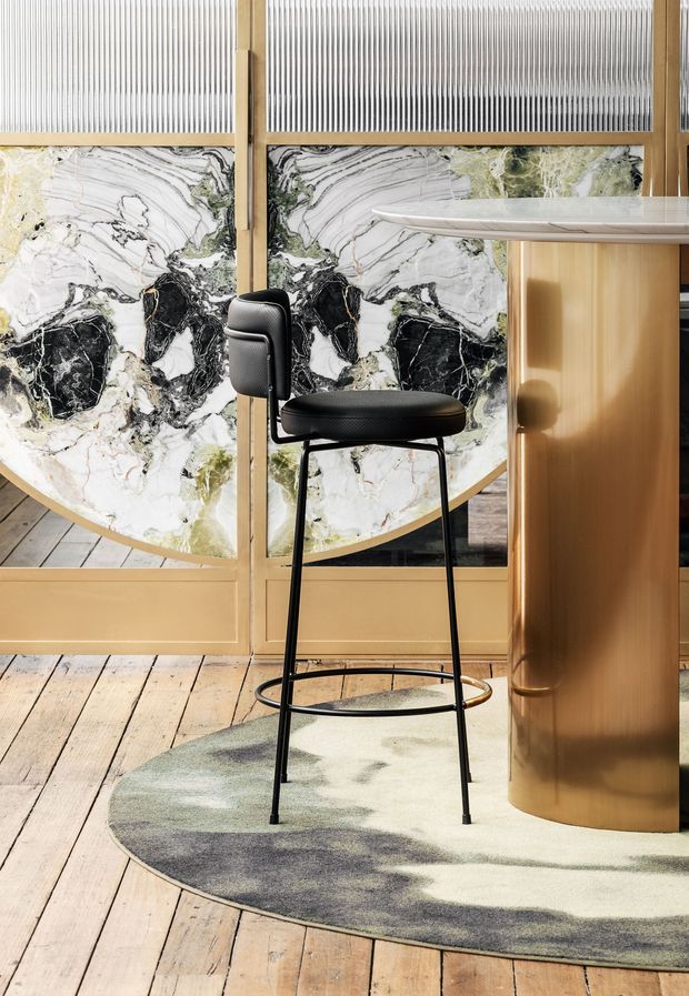 A refined interior vignette featuring a black bar stool beside a rounded bronze-toned counter or column. Behind it, striking veined marble panels are framed with warm timber and translucent ribbed glass. The space has pale timber plank flooring and a muted abstract rug, with strong sculptural geometry and layered natural materials.