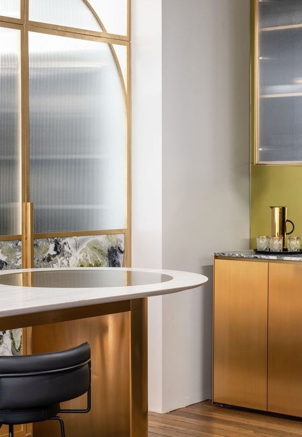 A refined contemporary dining and bar area featuring a curved white stone tabletop with an inset dark glass centre, warm timber and brass-toned cabinetry, fluted translucent glass panels in timber frames, and a compact drinks cabinet with a stone benchtop. A black upholstered bar stool and timber flooring add contrast.