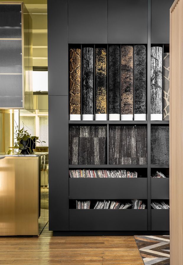 A close-up interior view of a sophisticated built-in storage and display wall. Matte black cabinetry with open shelving holds rolled textiles, fabric samples, and magazines, while a warm brass-toned cabinet and shelving unit sit to the left beside a window and indoor plant. Timber flooring, a patterned rug, and light timber wall panelling add warmth and contrast to the dark joinery.
