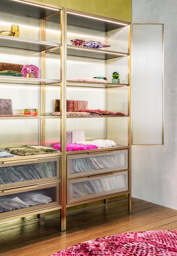 A richly styled walk-in wardrobe or dressing-room storage wall with floor-to-ceiling open shelving, translucent ribbed drawer fronts, integrated warm LED lighting, and brass-toned framing. Folded textiles, colourful accessories, and decorative objects are displayed on the shelves. The room has textured green and grey walls, timber floorboards, and a vivid pink patterned rug in the foreground.