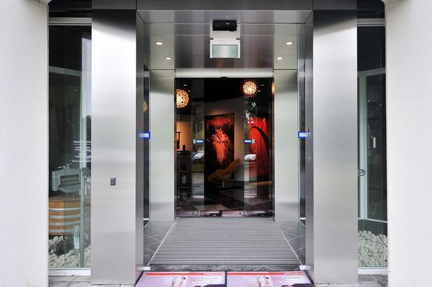 A modern elevator lobby or entry passage framed by polished stainless-steel wall panels and glazed doors. The central elevator entrance has a ribbed grey floor mat, reflective surfaces, recessed ceiling lighting, and dramatic red-and-black artwork visible inside. Decorative white stones and partial furnishings are visible through the side glazing.