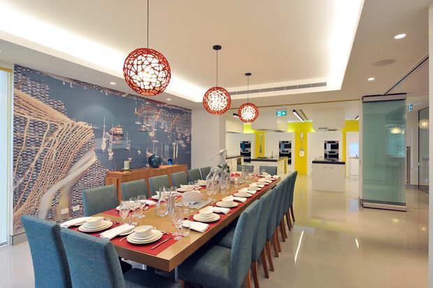 A contemporary dining area with a long timber dining table set for a large group, surrounded by blue upholstered chairs. Three red spherical pendant lights hang above the table, while a large aerial harbour or waterfront mural forms a feature wall behind a timber sideboard. The space opens toward a bright white kitchen with vivid yellow accents and integrated appliances. Glossy light flooring, recessed ceiling lighting, and glass partitions create a polished, modern commercial or hospitality-like interior.