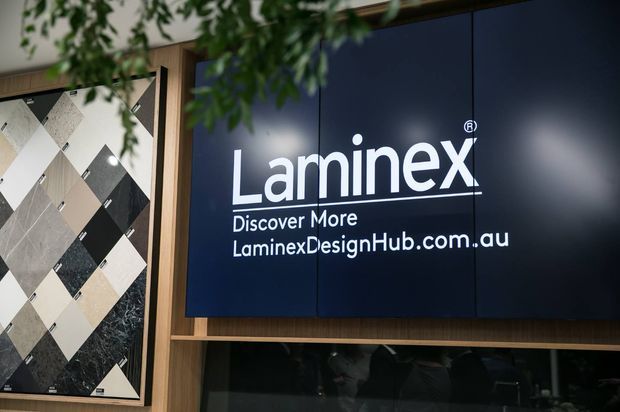 Close-up of a Laminex Design Hub display, featuring a large dark navy Laminex logo panel with white lettering, framed in timber-toned joinery. A tiled material sample board is visible on the left, with leafy greenery partially obscuring the upper foreground.