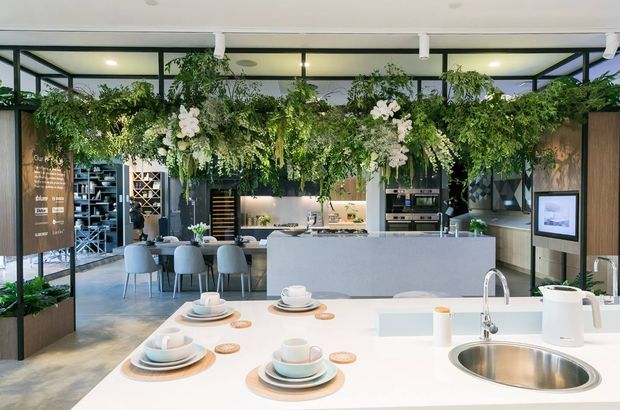 A bright contemporary kitchen showroom or hospitality-style kitchen display with a large white island and integrated round stainless-steel sink in the foreground. The space features dark-framed internal glazing, extensive timber cabinetry, a secondary kitchen island, dining stools, shelving, appliances, and abundant hanging greenery and white flowers overhead.