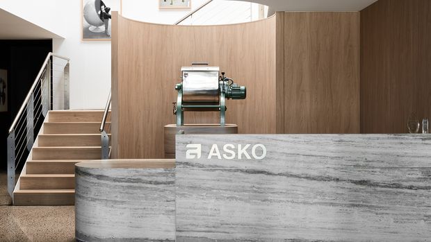 A contemporary commercial or hospitality-style interior featuring a large curved stone reception counter with an ASKO logo, a wood-panelled curved backdrop, and an open stairway with slim metal balustrading. A vintage-looking green and metallic machine is displayed behind the counter, with a skylight and framed artwork adding detail to the refined space.