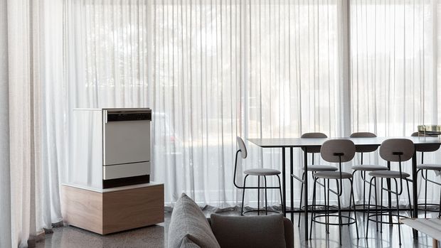 Bright contemporary open-plan interior with sheer floor-to-ceiling curtains, a compact wood-based kitchen or service cabinet, a high dining table with multiple stools, and the edge of a sofa in the foreground. The room has a clean, restrained palette and abundant filtered daylight.