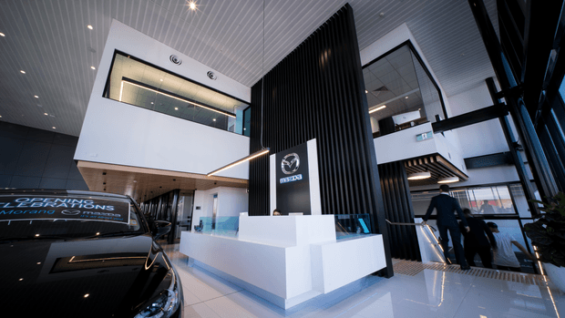 A bright, double-height commercial reception and showroom interior with a prominent Mazda-branded reception desk and tall black vertical feature screen. White architectural volumes, glossy light flooring, linear white ceiling panels, expansive glazing, mezzanine-level openings, and a stair descending to the right create a modern automotive dealership environment. A black vehicle occupies the foreground.