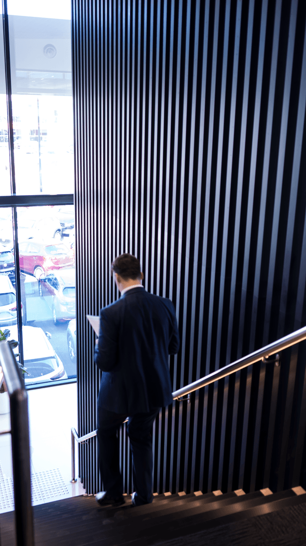 A dramatic interior view looking down a modern stairway beside a tall floor-to-ceiling window. The stair wall is clad in closely spaced vertical black slats, contrasted with a slim warm timber handrail and dark timber stair treads. A suited person descends the stairs, while parked cars are visible outside through the glazing. The image has a strong contemporary commercial or institutional character with high contrast and vertical rhythm.