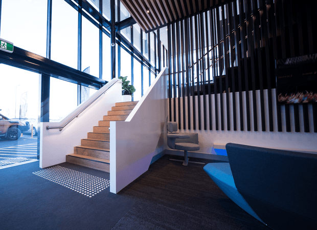 Contemporary commercial interior lobby with a stairway leading to an upper level. The space features a dramatic double-height glazed facade, dark vertical timber slats, a slatted ceiling, light-painted stair walls, timber stair treads, dark carpet flooring, and built-in or fixed lounge seating. An exit door and a small amount of exterior parking are visible through the glazing.