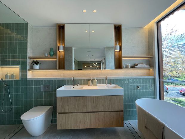 Contemporary bathroom with a double vanity, wall-mounted timber-look cabinetry, large central mirror, integrated open shelving, green tiled walls, and a freestanding bathtub beside full-height glazing. A wall-hung toilet and shower area are also visible.