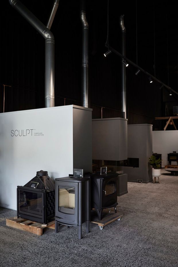A dark, industrial-style showroom displaying freestanding wood-burning stoves and fireplaces on timber pallets. Tall black flues rise through the high ceiling, with black track lighting, white display partitions, polished concrete flooring, and sparse product displays. The wall is branded “SCULPT Fireplace Collection.”