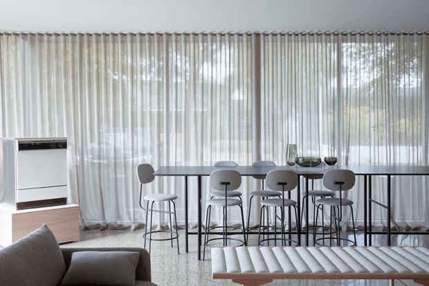 Bright contemporary open-plan living and dining area with floor-to-ceiling sheer curtains, a long high dining table with bar stools, a sculptural upholstered bench in the foreground, and a freestanding fireplace set into low timber cabinetry. The restrained palette of whites, greys, and light timber creates an airy, refined interior.