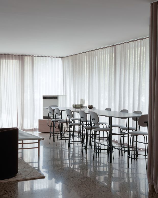 Bright, minimalist dining area with a long dark dining table surrounded by numerous light upholstered chairs with slender black metal frames. Floor-to-ceiling sheer curtains wrap around the corner glazing, creating a softly lit space. The polished speckled floor reflects the furniture and daylight, while a compact sideboard or cabinet and a partial lounge seating area are visible at the edges.