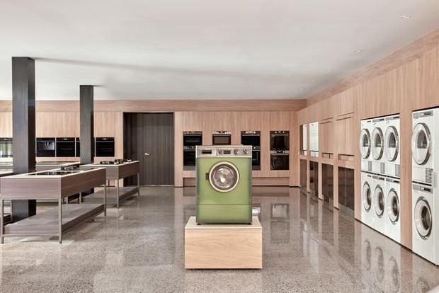 A spacious contemporary laundry showroom or utility space with extensive pale timber cabinetry, integrated black appliances, a central vintage-style green washing machine displayed on a light timber plinth, and multiple white front-loading washers and dryers along the right wall. The polished terrazzo-like floor reflects the cabinetry and appliances, while dark structural columns and a clean white ceiling add contrast.