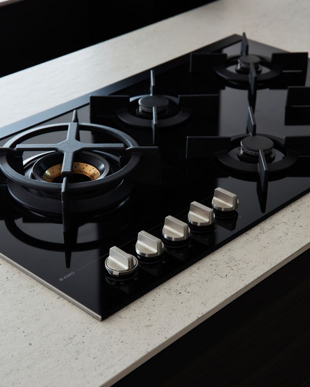 Close-up product-style view of a black glass gas cooktop with four burners, cast-iron pan supports, and metallic control knobs, set into a light speckled stone benchtop. Dark cabinetry is visible beneath the counter.