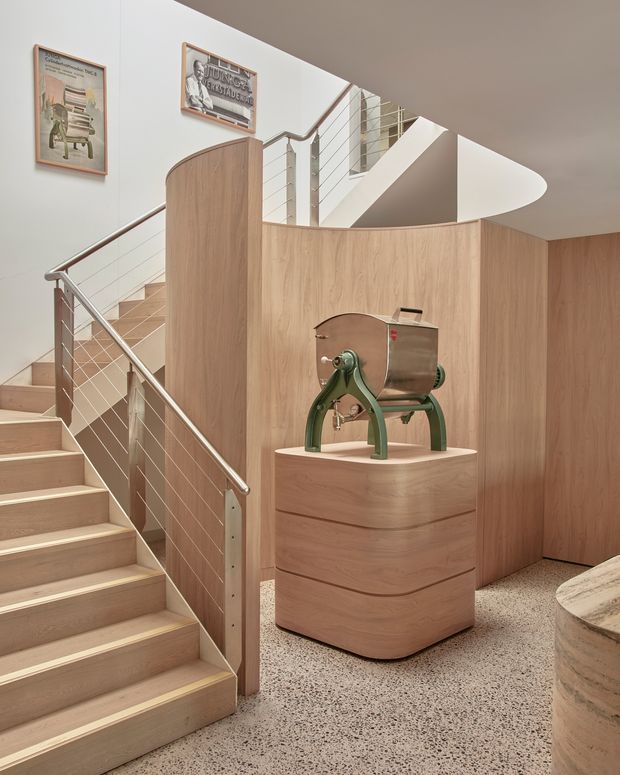 Interior view of a sculptural timber-lined stairway and landing, featuring pale wood stair treads, curved timber partition walls, stainless-steel cable balustrades, and a terrazzo floor. A vintage-style green and stainless-steel coffee machine sits on a rounded timber plinth, while framed posters decorate the white wall. The stair rises beneath a curved white ceiling and continues to an upper level.