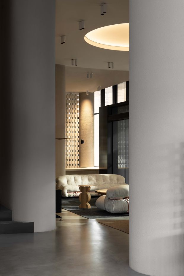 A refined contemporary interior viewed through a curved opening, featuring a sunken lounge with a cream tufted sofa, rounded ottomans, sculptural wood coffee table, dark geometric rug, large black-framed glazing, and a patterned breeze-block screen. The space has polished concrete flooring, curved plaster columns, high ceilings, recessed and concealed lighting, and a warm neutral palette.