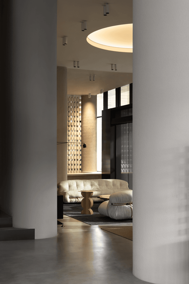A refined contemporary living room viewed through a narrow opening between curved plaster columns. The space features a sculptural cream sofa and rounded ottoman, a dark wood coffee table, polished concrete flooring, tall black-framed glazing, a textured feature wall with patterned blockwork, and a large recessed oval ceiling light. Warm natural light and soft neutral tones create an elegant, calm atmosphere.