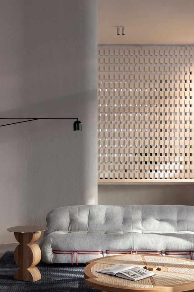 A refined living room with a large tufted light-grey sofa, sculptural wooden side table, round woven coffee table, black articulated wall lamp, textured pale walls, and a distinctive perforated or lattice-like cream room divider. The space is softly lit with a warm, neutral palette and strong geometric detailing.