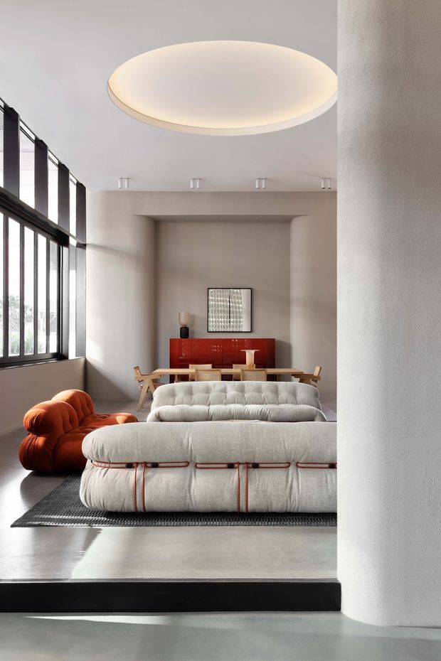 A refined open-plan living and dining space with a large upholstered modular sofa in the foreground, an orange sculptural lounge chair, a wooden dining table and chairs, and a red sideboard at the rear. The room has pale textured walls, polished concrete flooring, tall black-framed windows, and a large circular pendant light. The composition is minimalist yet expressive, with strong geometric forms and warm accent furniture.