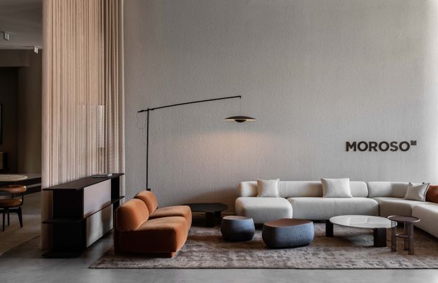 A refined contemporary living room and adjacent open kitchen/dining area. The space features a large cream sectional sofa, sculptural brown leather lounge chair, low rounded coffee tables, a textured area rug, a slender black floor lamp, and a vertically ribbed timber partition screening the kitchen. A textured light-grey wall displays the MOROSO logo.