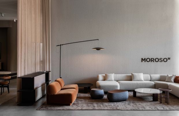 A refined contemporary living room or showroom interior featuring a large cream sectional sofa, sculptural brown leather lounge chair, low rounded coffee tables, and a large neutral area rug. A slim black floor lamp and textured light wall create a minimalist backdrop, with vertical timber screening and dark built-in cabinetry visible to the left. The MOROSO wall logo suggests a furniture showroom or branded display space.