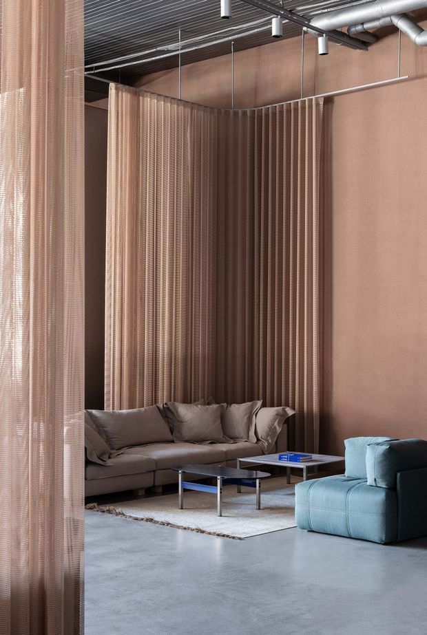 A contemporary lounge area with a muted taupe and grey palette. The space features sculptural sofas and armchairs arranged around low rectangular coffee tables on a pale rug. Floor-to-ceiling ribbed translucent curtains create a partition and textured backdrop, while exposed metal ductwork, suspended cylindrical lights, and a dark linear ceiling add an industrial character. The floor appears to be smooth polished concrete.
