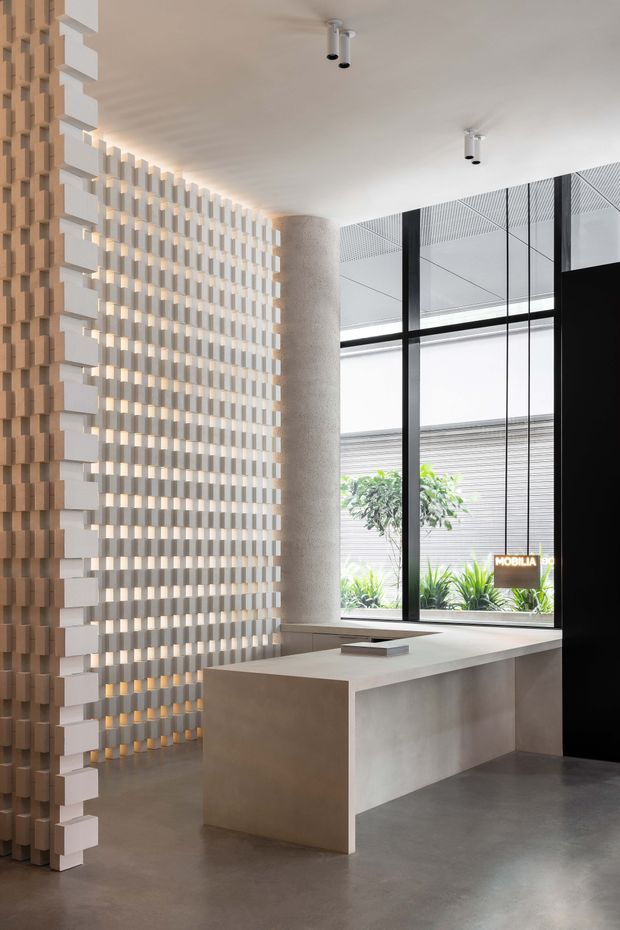 A refined contemporary commercial interior featuring a sculptural textured feature wall of repeating light timber-toned blocks, a cylindrical textured column, a minimalist concrete or stone reception counter, polished grey flooring, and full-height black-framed glazing. The space is bright, uncluttered, and carefully detailed, with planted landscaping visible outside.