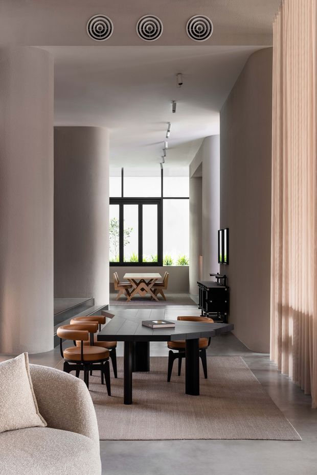 A refined open-plan interior with a dining and living area. The space features a dark sculptural dining table with curved wooden chairs, a pale upholstered sofa in the foreground, smooth light-toned walls, a curved column, concrete-look flooring, a large neutral rug, sheer vertical curtains, and expansive black-framed glazing. Track lighting and circular ceiling vents contribute to the clean, contemporary design.
