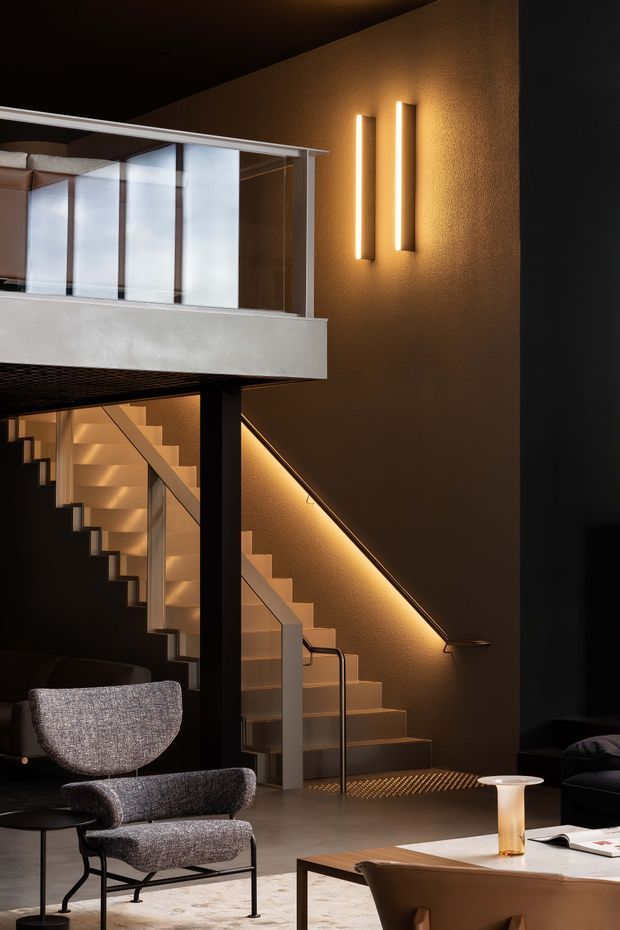 A dramatic contemporary interior featuring a double-height stair hall and mezzanine. A sculptural staircase with illuminated treads and a warm linear handrail light rises beside a textured dark wall, while a glazed mezzanine with a pale concrete fascia overlooks the space. The lower level includes a refined lounge area with sculptural upholstered armchairs, a dark sofa, a timber coffee table and warm accent lighting.