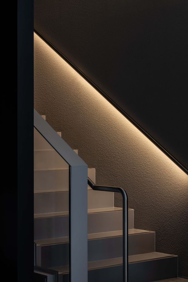 Close-up architectural photograph of a modern stairway with dark textured walls, warm concealed LED strip lighting following the stair soffit, pale stair treads, and a dark metal handrail with a solid side balustrade. The composition emphasizes the geometric stair profile and dramatic ambient lighting.