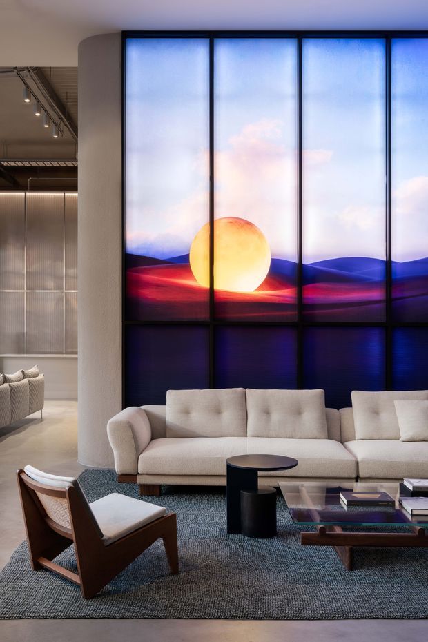 A sophisticated contemporary lounge with a large cream sectional sofa, sculptural dark coffee tables, mid-century-inspired leather lounge chairs, and a textured grey rug. A dramatic floor-to-ceiling illuminated graphic feature wall depicts a glowing sun over layered blue and magenta landscapes, framed in black. The room also includes a curved pale column, exposed ceiling services and track lighting, and translucent screening visible to the side.