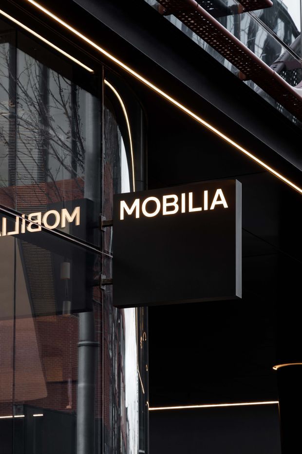 Close-up exterior view of a contemporary commercial storefront or building entrance, featuring a black projecting sign with illuminated “MOBILIA” lettering, dark metal architectural surfaces, glass facade, integrated linear lighting, and curved detailing. Reflections of surrounding urban buildings are visible in the glazing.