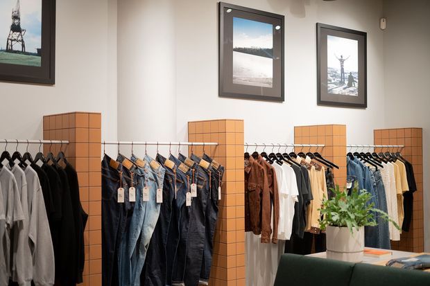 A contemporary clothing retail showroom with neatly arranged garments hanging from wall-mounted rails. Warm terracotta tiled display partitions divide sections for jackets, shirts, jeans and other apparel, while framed photographic artwork and a potted plant add decorative detail. A dark upholstered seating area is partially visible in the foreground.
