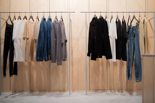 A minimalist clothing display area with two freestanding metal garment racks arranged against pale timber wall panelling. Neutral-toned shirts, jackets, sweaters, trousers and jeans hang neatly on wooden and metal hangers. The space has a light patterned tile floor and a clean, retail-like presentation.