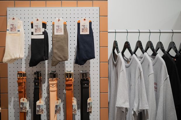 Retail clothing display featuring socks and belts mounted on a white pegboard against an orange brick wall, alongside a metal garment rail holding neatly arranged grey and black T-shirts on hangers.