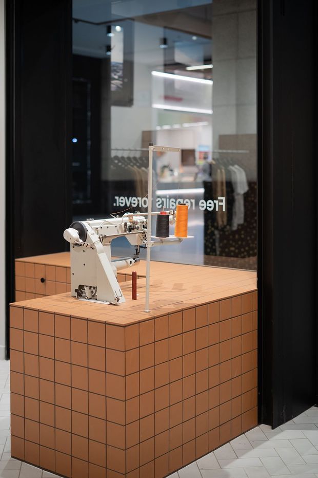 A close-up view through a dark-framed glass storefront into a contemporary clothing retail space. A vintage-style industrial sewing machine with orange and dark thread sits on a terracotta square-tiled display counter, with clothing racks and illuminated shelving visible behind it. The floor is finished with pale geometric tiles, and reversed storefront lettering appears on the glass.