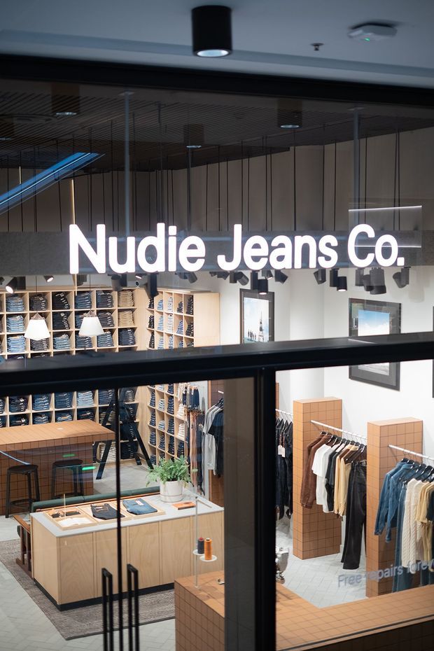 Interior view of a modern Nudie Jeans Co. retail clothing store, photographed through glass doors or a storefront partition. The shop features extensive open shelving filled with folded denim, clothing racks, pale timber display counters, terracotta-toned tiled display plinths, white patterned floor tiles, suspended pendant lighting, and a dark linear ceiling treatment. A large illuminated Nudie Jeans Co. sign dominates the upper portion of the image.