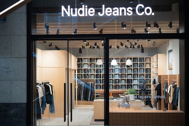Front-facing view through the glazed entrance of a Nudie Jeans Co. retail store. The commercial interior features pale timber wall panelling and shelving, denim merchandise displays, black track lighting, pendant lights, a central timber retail counter, and dark-framed full-height glazing and doors.
