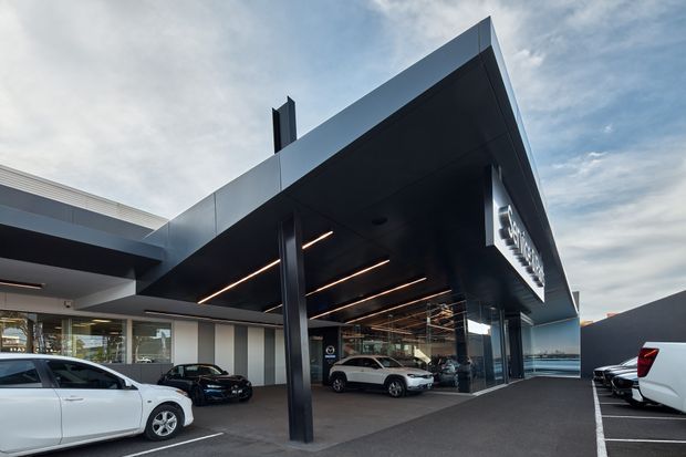 Exterior view of a contemporary automotive dealership with a large cantilevered dark canopy, glazed showroom frontage, integrated linear lighting, and vehicles displayed beneath the entry structure. The building uses crisp geometric forms, dark metal cladding, extensive glazing, and a paved forecourt.