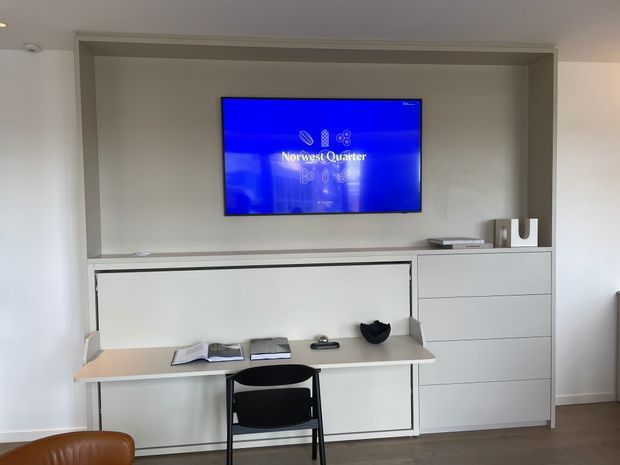 A clean, minimalist built-in media and workspace wall featuring a wall-mounted television within a recessed niche, integrated white cabinetry, a compact desk surface, and a black chair. Neutral painted walls and dark timber flooring create a simple, functional interior.